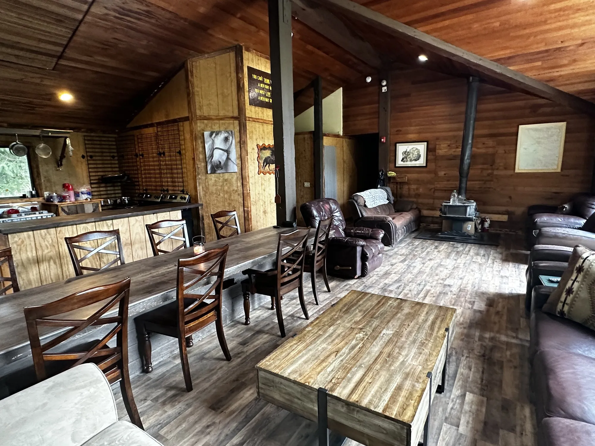 The great room — a long dining table, leather recliners, and a wood stove under a vaulted cedar ceiling